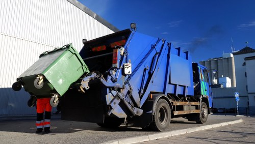 A large blue refuse collection truck is parked on a paved surface alongside a white industrial building under a clear blue sky. The truck's rear compactor mechanism is in the process of emptying a green wheeled bin, which is tilted and positioned at the back of the vehicle. The green bin appears to be made of durable plastic with a textured surface and four small caster wheels at its base. A worker wearing a high-visibility orange safety vest and dark trousers is standing near the bin, partially obscured, observing the loading process. The scene is set outdoors, likely in an industrial or commercial area, with the truck aligned parallel to the building and adjacent to a curb. This image visually illustrates rubbish collection or waste disposal services provided by Commercial Waste Seven Kings, emphasizing professional and efficient waste removal in the Seven Kings area, within the postal code vicinity.