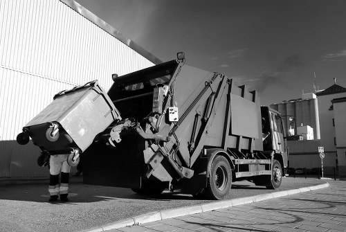A large black and white rubbish collection truck with an open rear loading mechanism is positioned on a paved street near a white corrugated metal building, with a worker standing nearby. The worker, dressed in work trousers and a high-visibility vest, is emptying a large, rectangular, light-colored bin into the rear hopper of the truck. The bin appears to be made of plastic and has a textured surface, with wheels visible at the bottom. The truck’s body features mechanical arms and hydraulic components designed for lifting and emptying waste bins, with the scene set in an urban or industrial area. In the background, there are industrial structures including storage tanks or silos, and the sky is partly cloudy. This scene illustrates a commercial waste collection operation consistent with rubbish removal services offered by Commercial Waste Seven Kings, serving areas in and around Seven Kings, Essex.