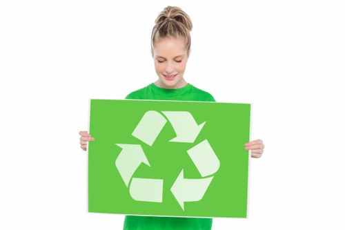 A young woman with light brown hair tied back in a ponytail, wearing a bright green long-sleeved top, is smiling gently while holding a large, rectangular green sign featuring a white recycling symbol composed of three chasing arrows forming a triangle. She stands against a plain white background, with her hands gripping the sides of the sign at chest level. The image emphasizes environmental awareness, recycling, and waste management themes relevant to rubbish removal services in Seven Kings, London, highlighting eco-friendly practices associated with commercial waste solutions. The woman's approachable expression and the clear recycling icon reinforce the message of sustainability in waste disposal efforts.