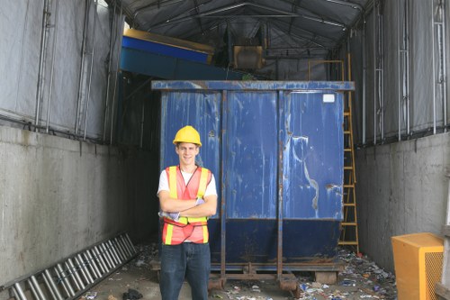 A young man wearing a yellow safety helmet and a high-visibility vest stands with arms crossed in front of a large, blue industrial skip situated inside a metal-walled storage area or warehouse. The skip, made of sturdy metal with visible scratches and wear marks, is positioned on a concrete floor cluttered with small bits of rubbish and debris. To the right of the skip, there is a yellow waste container or box, and behind it, a yellow ladder leaning against the back wall. The surrounding environment appears industrial, with metallic walls and roofing panels, and the area is lit with natural and artificial light. The scene relates to waste management services in the Seven Kings region, exemplifying site preparation or rubbish collection operations managed by Commercial Waste Seven Kings.