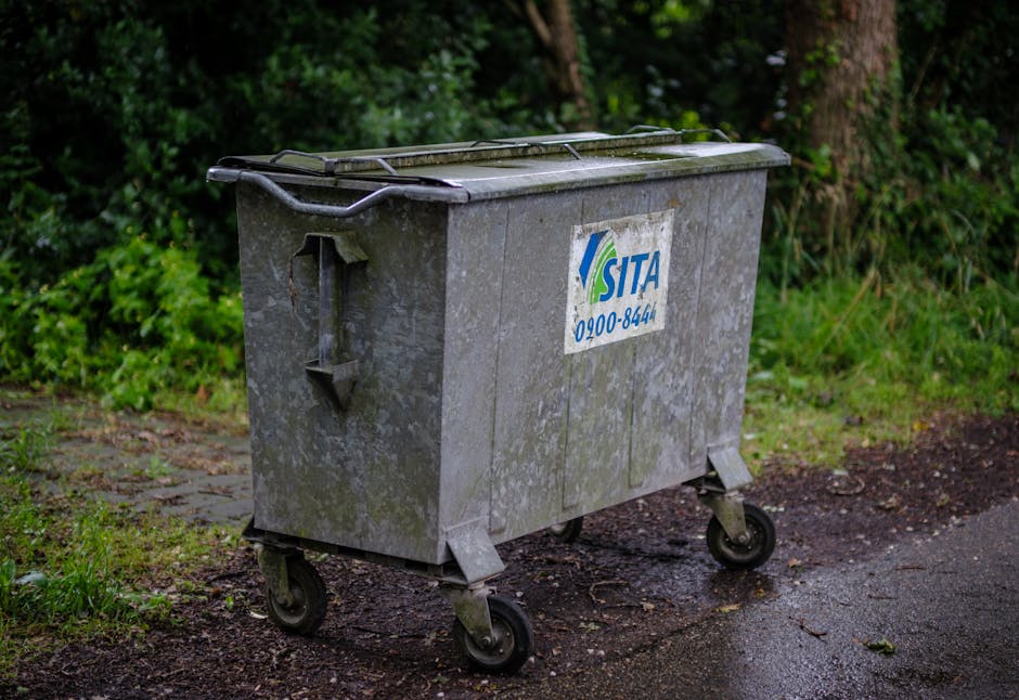 A large, rectangular metal wheelie bin with a slightly weathered and dirty surface sits on a damp, uneven asphalt pathway. The bin is positioned in an outdoor area, with lush green foliage and trees i