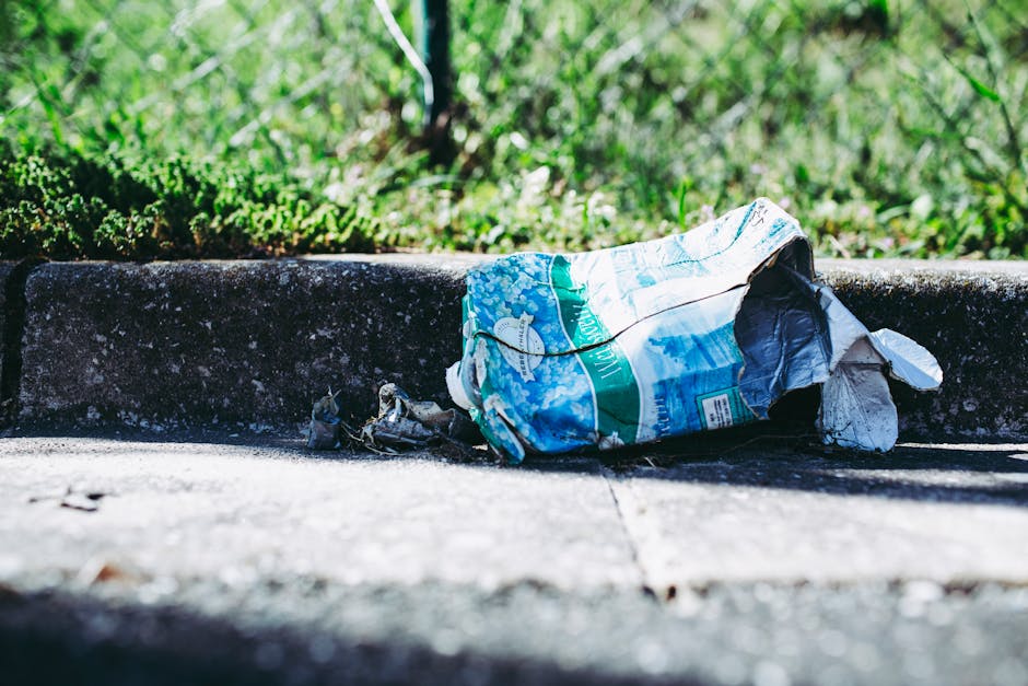 A large, rectangular metal wheelie bin with a slightly weathered and dirty surface sits on a damp, uneven asphalt pathway. The bin is positioned in an outdoor area, with lush green foliage and trees i