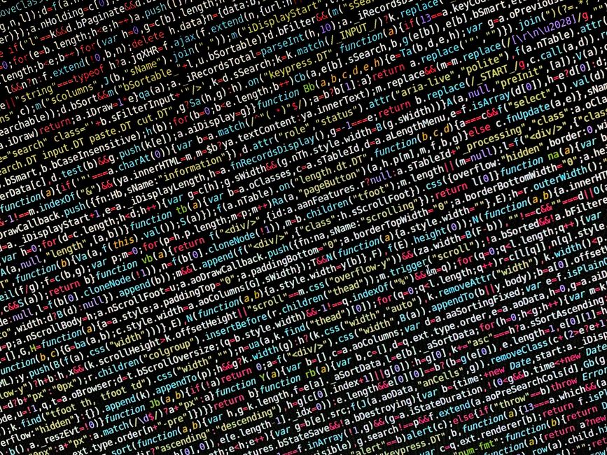A photograph of a cluttered computer screen displaying lines of colourful computer code in various shades of black, red, green, yellow, blue, and white. The code consists of text, symbols, and numbers